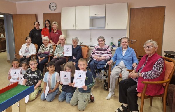 Auf dem Bild sieht man eine Gruppe Seniorinnen. Vor ihnen knien mehrere Kinder, die stolz ihre Kegelurkunden in die Kamera halten und fröhlich lachen.