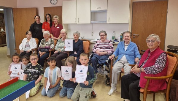 Auf dem Bild sieht man eine Gruppe Seniorinnen. Vor ihnen knien mehrere Kinder, die stolz ihre Kegelurkunden in die Kamera halten und fröhlich lachen.