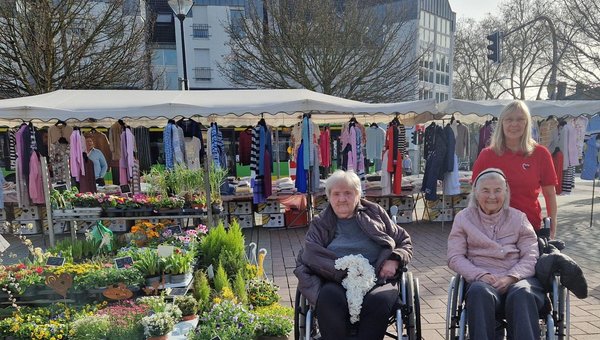 Zwei Seniorinnen im Rollstuhl besuchen den Markt. Sie sitzen neben einem bunten Blumenstand, während eine Frau hinter ihnen steht. Alle lächeln entspannt in die Kamera.
