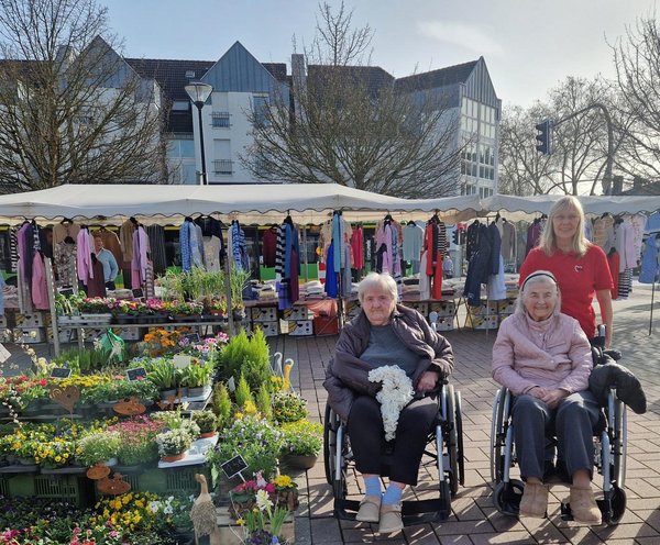 Zwei Seniorinnen im Rollstuhl besuchen den Markt. Sie sitzen neben einem bunten Blumenstand, während eine Frau hinter ihnen steht. Alle lächeln entspannt in die Kamera.