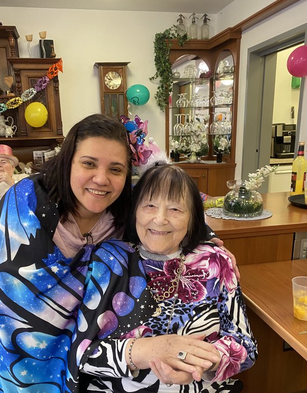 Zwei verkleidete Frauen lachen in die Kamera, eine Seniorin mit Krönchen wird von einer als Schmetterling verkleideten Frau im Arm gehalten