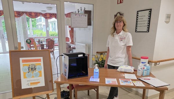 Eine Frau steht hinter einem Tisch mit Desinfektionsmitteln, Flyern und einem Aufsteller zum Internationalen Tag der Händehygiene.