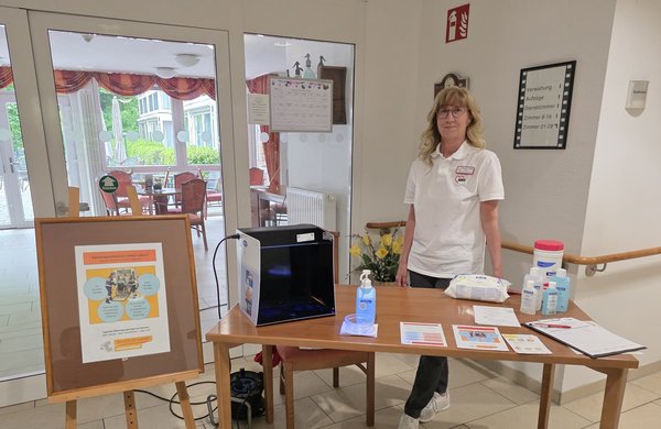 Eine Frau steht hinter einem Tisch mit Desinfektionsmitteln, Flyern und einem Aufsteller zum Internationalen Tag der Händehygiene.