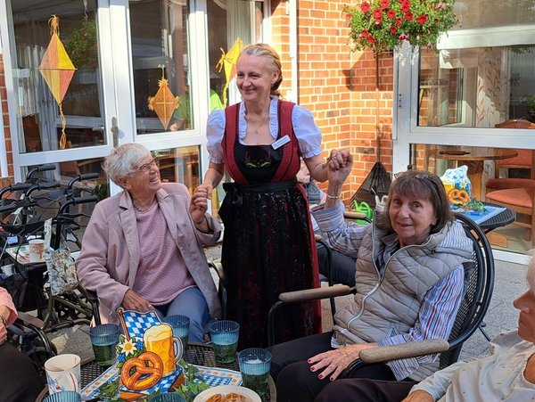 Eine Gruppe Seniorinnen sitzt an einem bayerisch geschmückten Tisch. Eine Frau im Dirndl steht zwischen ihnen und hält zwei Seniorinnen an den Händen.