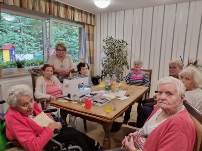 Auf dem Bild sieht man eine Gruppe Seniorinnen, die rund um einen Tisch sitzen, auf dem eine Nähmaschine steht. Alle lächeln freundlich in die Kamera.