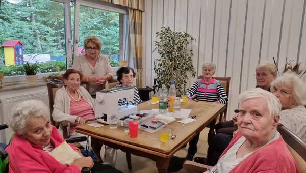 Auf dem Bild sieht man eine Gruppe Seniorinnen, die rund um einen Tisch sitzen, auf dem eine Nähmaschine steht. Alle lächeln freundlich in die Kamera.